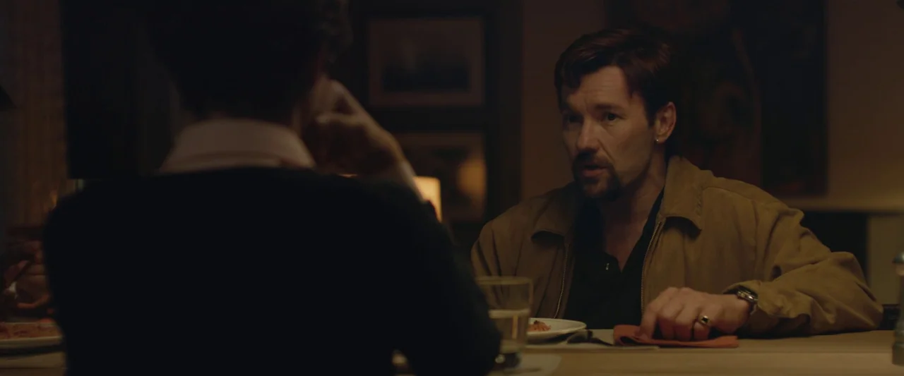 The Gift (2015) movie still: medium — A man sits at a wooden table in a dimly lit, residential dining room. He leans forward while listening to…