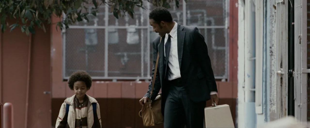 The Pursuit of Happyness (2006) movie still: fullbody — An urban sidewalk area features a chain link fence and a concrete wall backdrop. An adult…