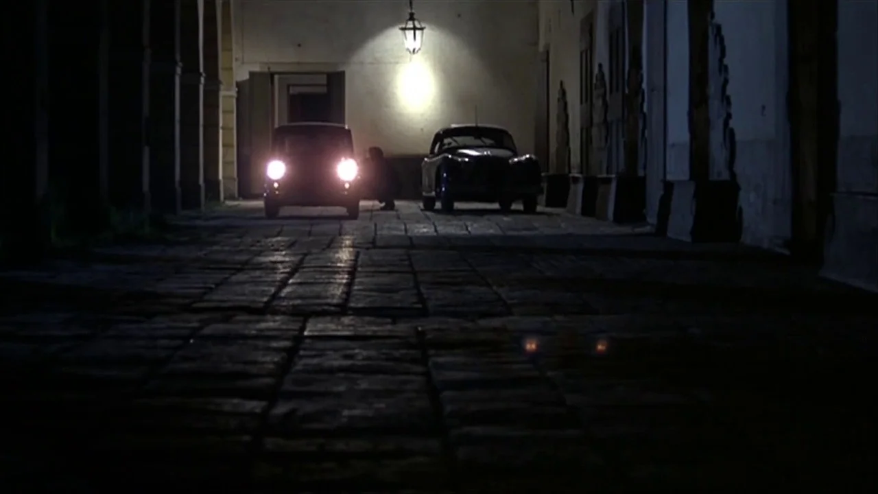 A Quiet Place In The Country (1968) movie still: wide — A dark cobblestone path leads toward an illuminated alcove featuring two parked vintage…