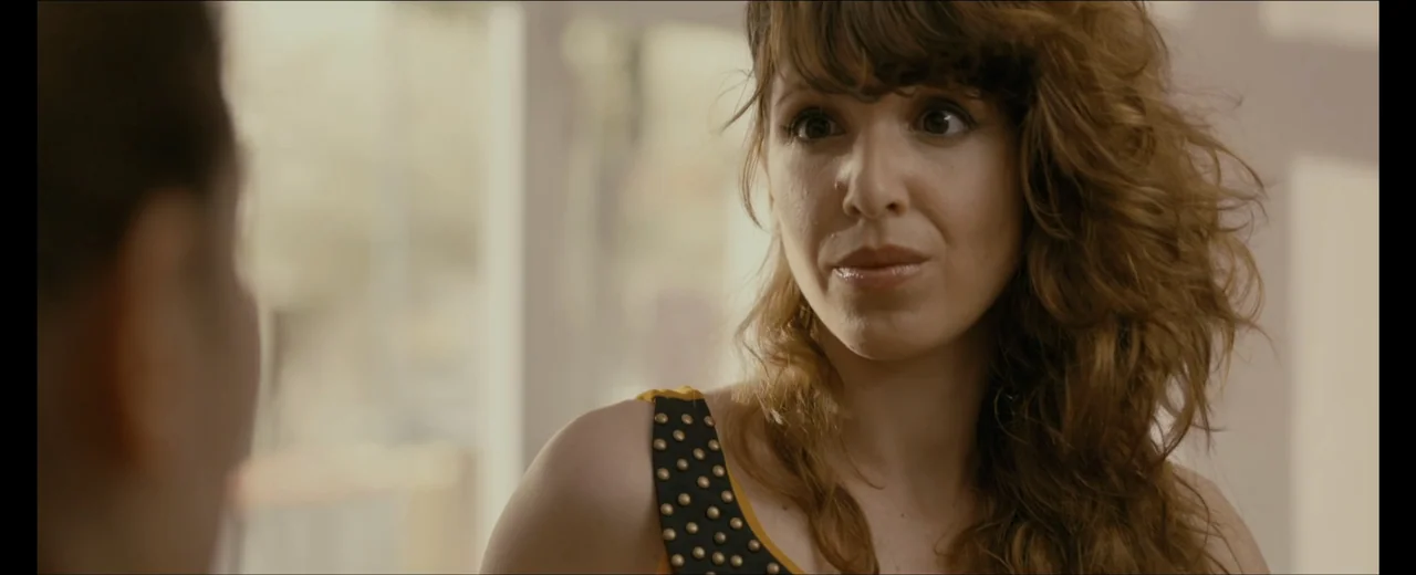 Faraday (2013) movie still: closeup — An interior space with bright light coming through a large window. A woman with long wavy hair faces an…