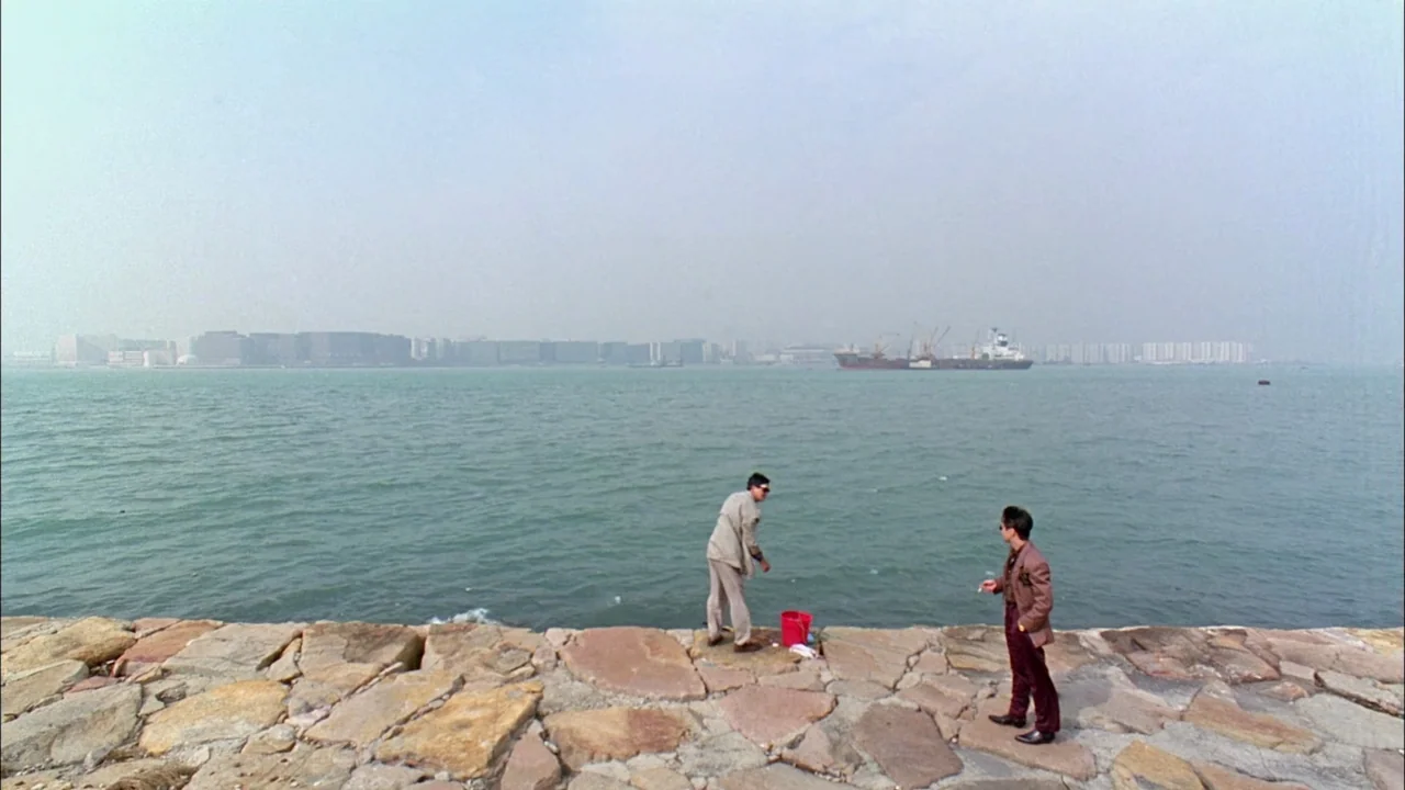 Hard Boiled (1992) movie still: wide — A stone paved waterfront edge overlooks a wide bay with a distant cargo ship. Two men stand on the uneven…