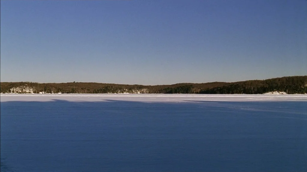 Away From Her (2006) movie still: establishing — A vast frozen lake stretches toward a low horizon line topped by bare trees. The foreground is cast…