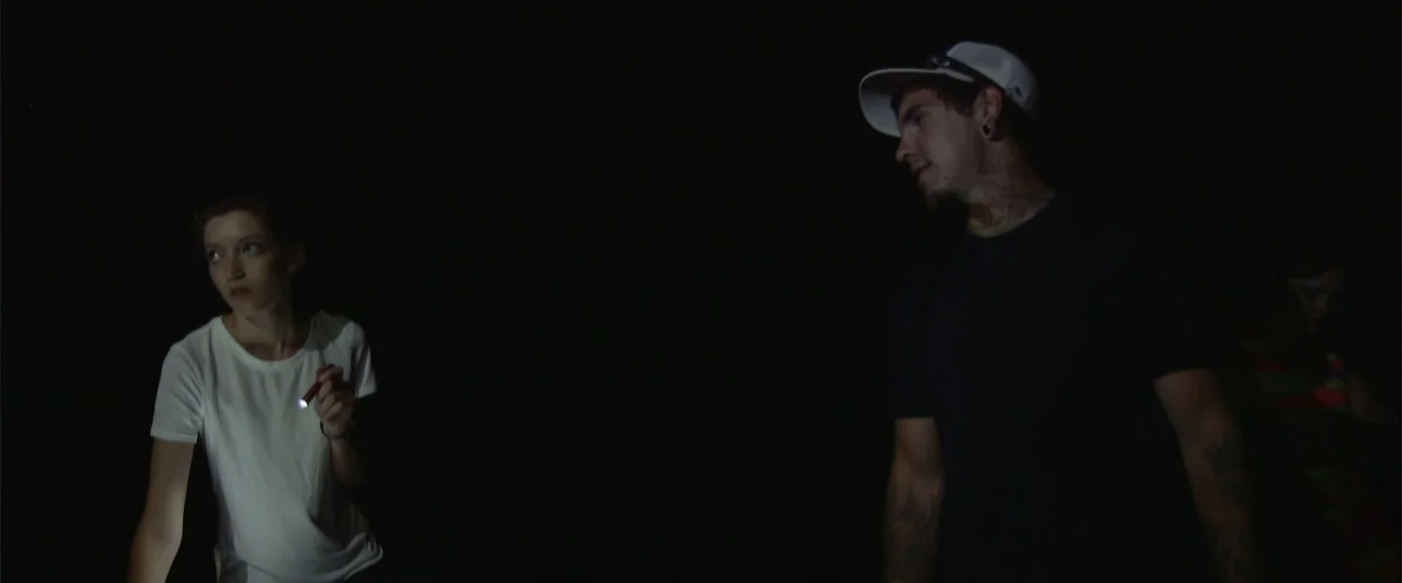 Darkness Waits (2020) movie still: medium — Two individuals stand in an almost completely dark environment illuminated by a small flashlight. A…