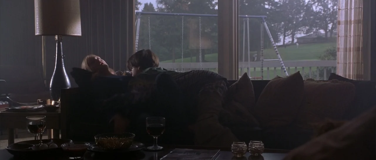 Bandits (2001) movie still: medium — A living room interior features a sofa positioned before a large window overlooking a lawn. A man and woman…
