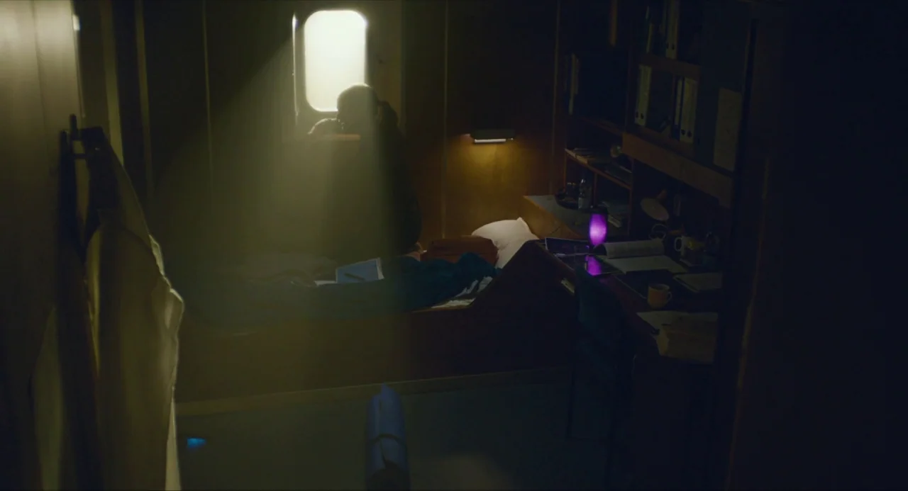 Submergence movie still: wide — A confined, wood-paneled cabin with a small window and integrated desk unit. A solitary person sits on the narrow…