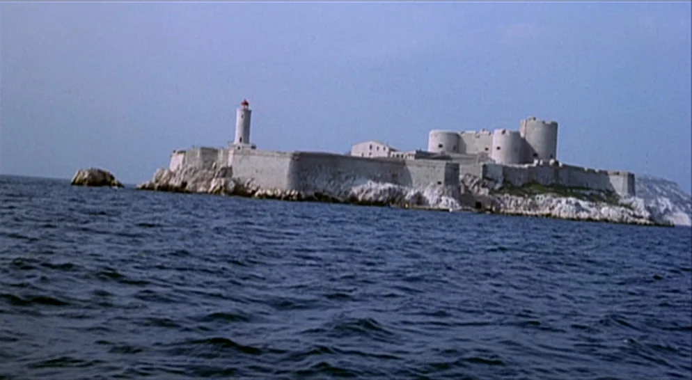 The French Connection (1971) movie still: establishing — A stone fortress sits atop a rocky island surrounded by deep blue sea water. A lighthouse…
