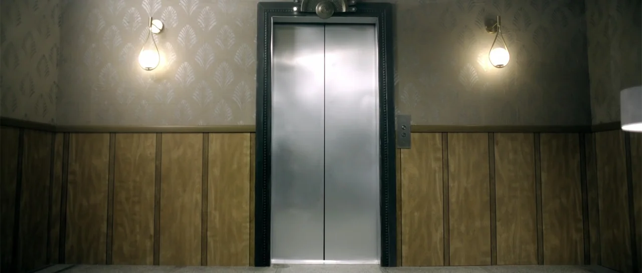 Elevator Game (2023) movie still: medium — A symmetrically composed elevator lobby features vertical wood wainscoting and ornate patterned beige…