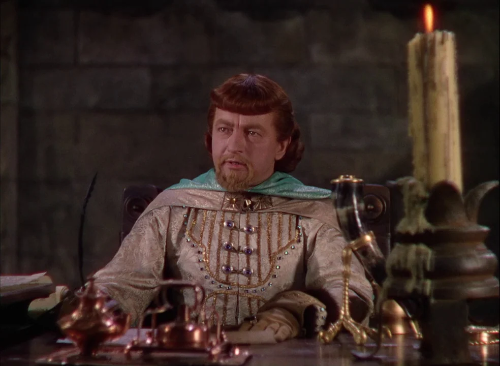 The Adventures of Robin Hood (1938) movie still: medium — A man in medieval finery sits at a wooden desk inside a stone chamber. He looks toward the…