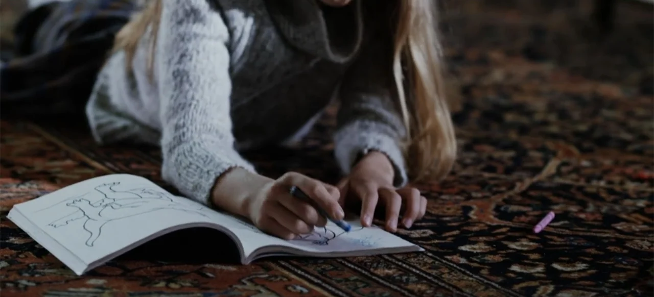 The Russian Bride (2019) movie still: medium — A child lies on a patterned carpeted surface while drawing in a book. The subject uses a blue crayon…