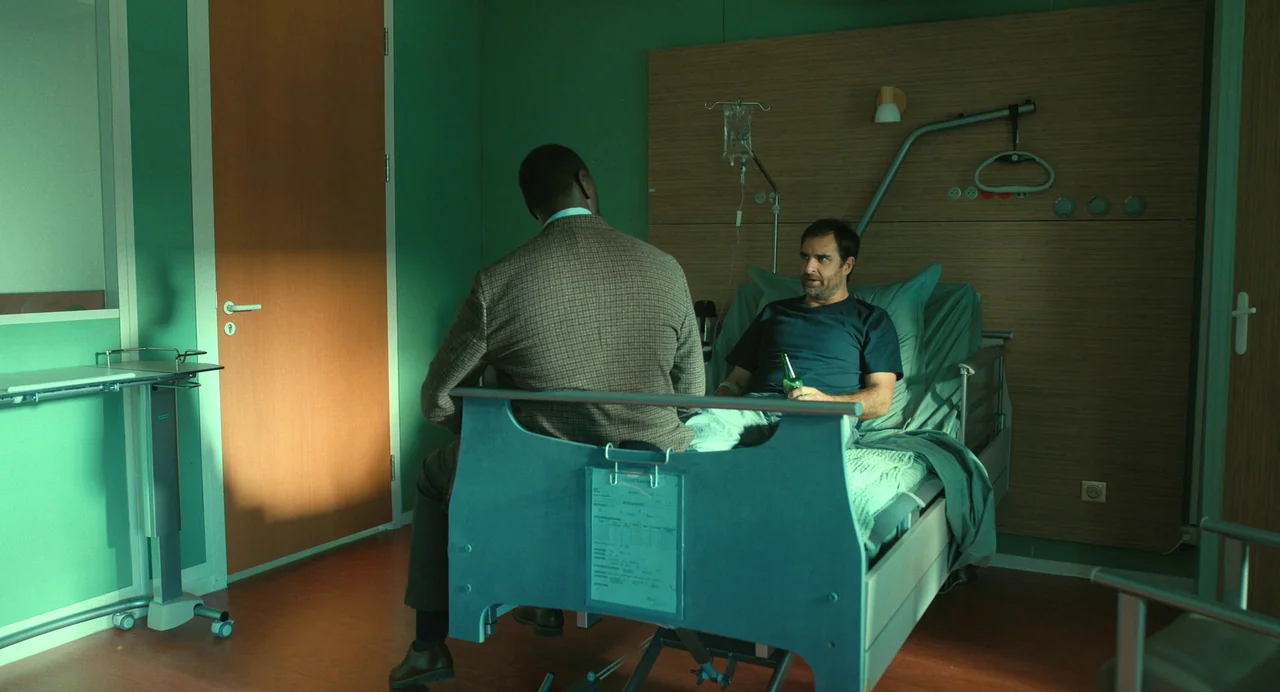The Killer (2024) (2024) movie still: medium — A sterile room with green walls and a wooden headboard features a modern bed. One man sits on the…