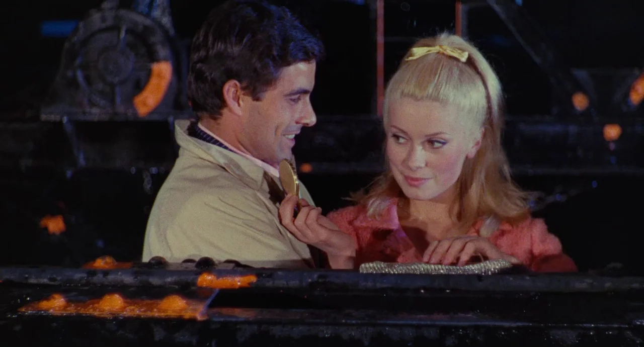 The Umbrellas Of Cherbourg (1964) movie still: medium — A dark industrial environment features massive black metal machinery with orange glowing…