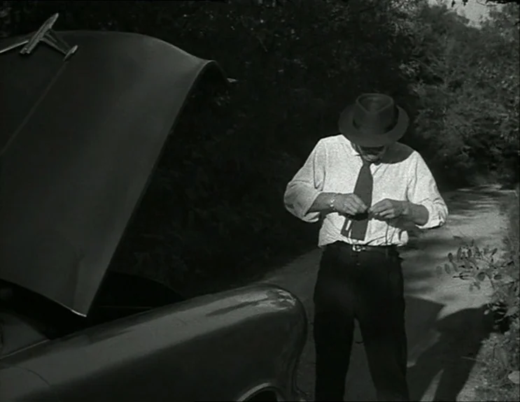 À Bout De Souffle (Breathless) (1960) movie still: medium — A man stands beside a parked car on a narrow, dirt country road. The hood of the car is…