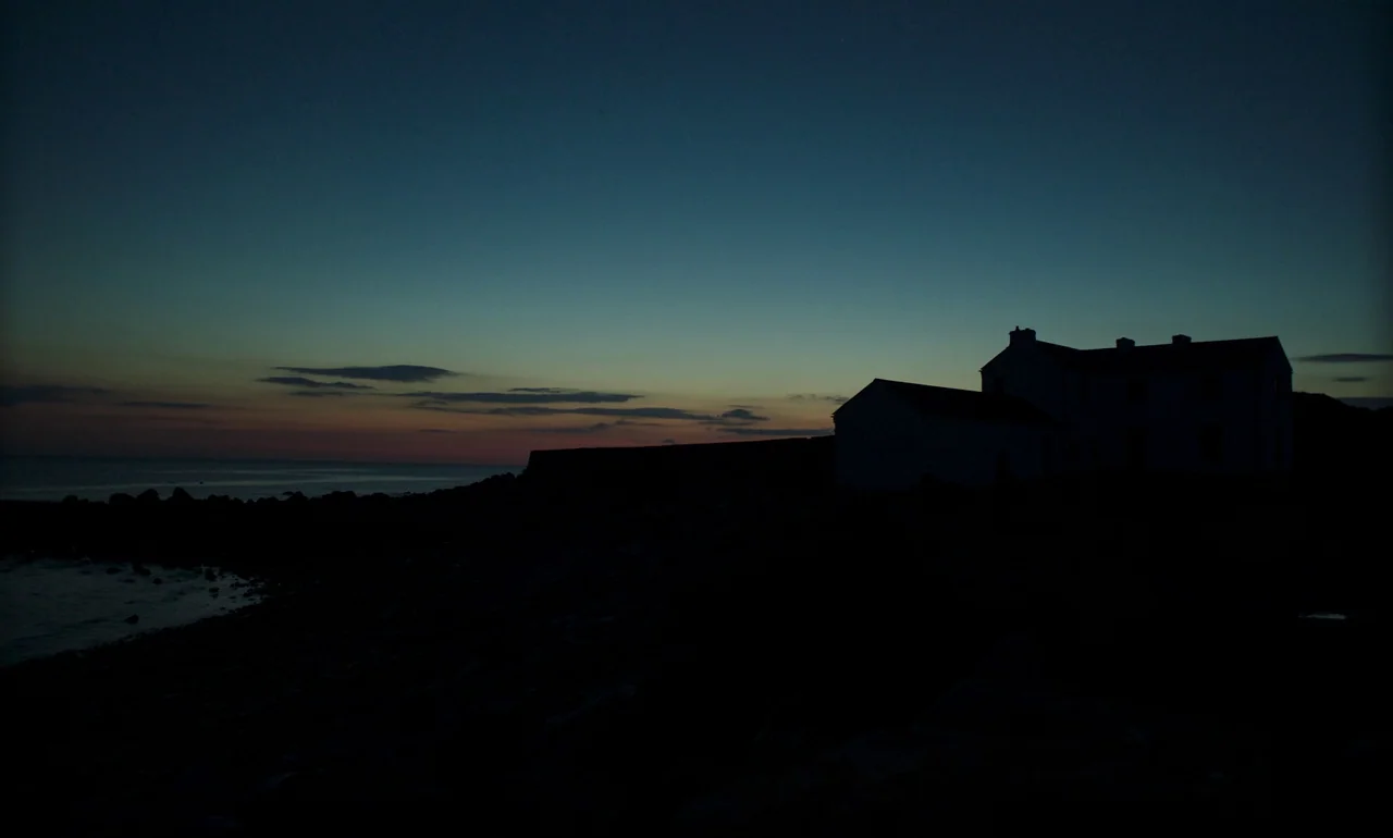 It Is In Us All (2022) movie still: establishing — A dark, silhouetted building stands near a rocky shoreline at the edge of water. The distant…
