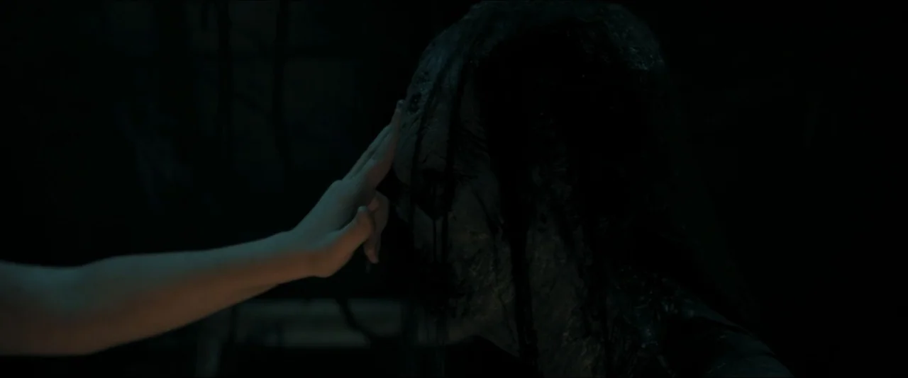 It Feeds (2025) movie still: closeup — A dark, underexposed environment with minimal visibility of the background architecture. A human hand extends…
