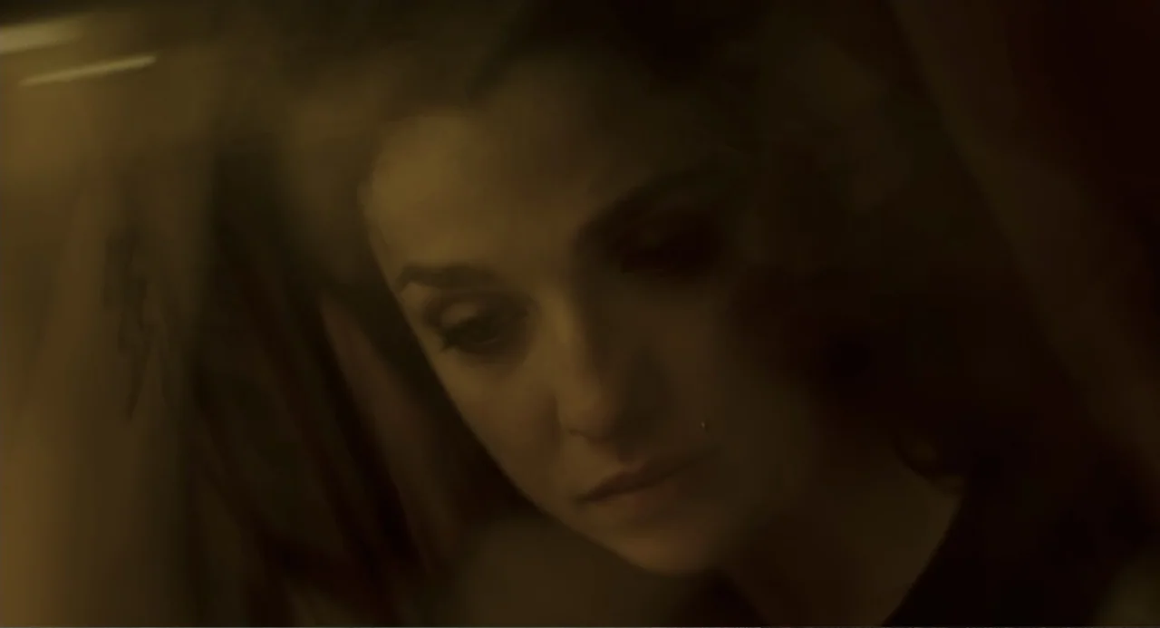 Back to Black (2024) movie still: closeup — A dimly lit interior space with brown, low-contrast shadows surrounding the subject. A woman looks down…