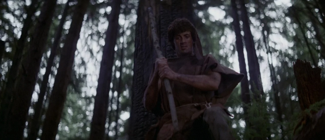 First Blood (1982) movie still: medium — A dense grove of tall, dark evergreen trees surrounds a man in a poncho. The man stands between two large…
