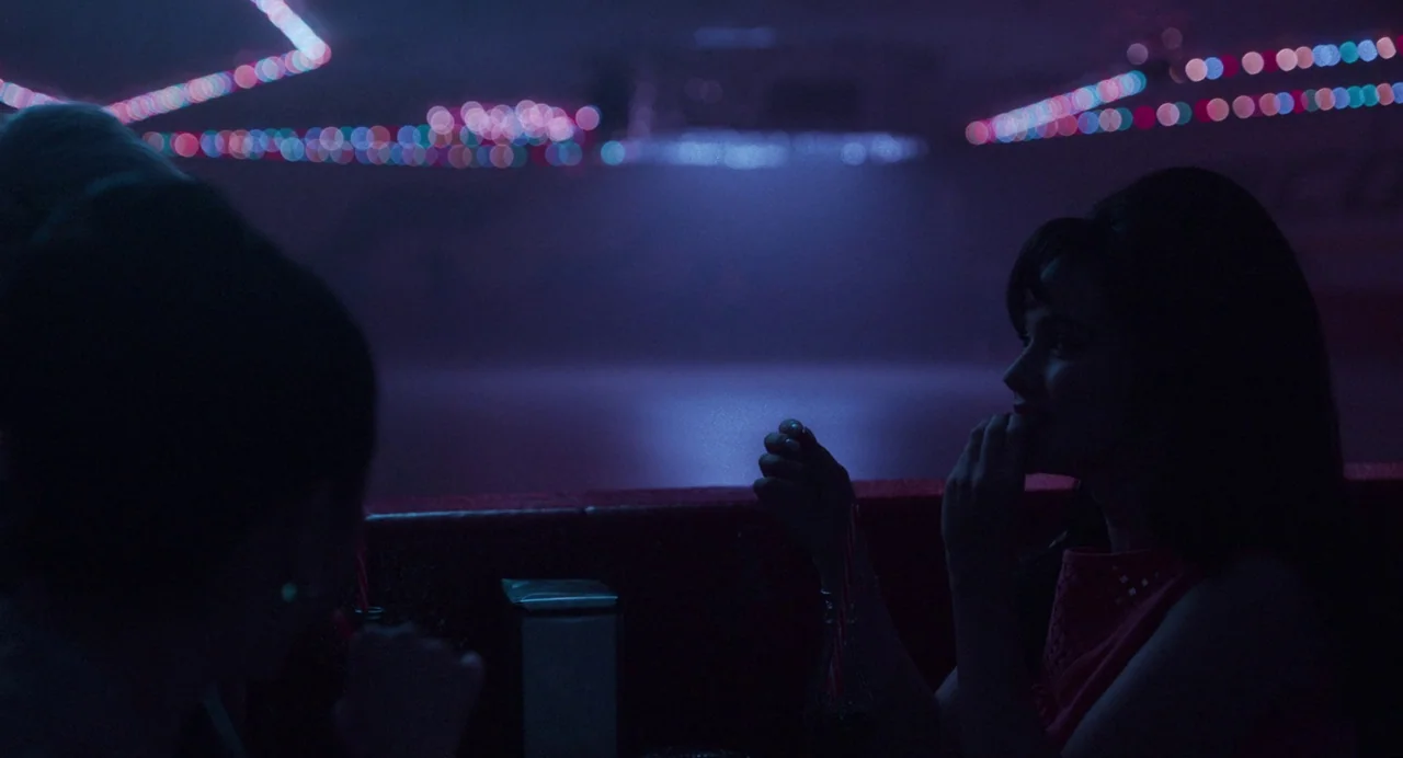 Priscilla (2023) movie still: medium — A dimly lit diner booth at night with soft blue and pink ambient light. Two women sit across from each other…