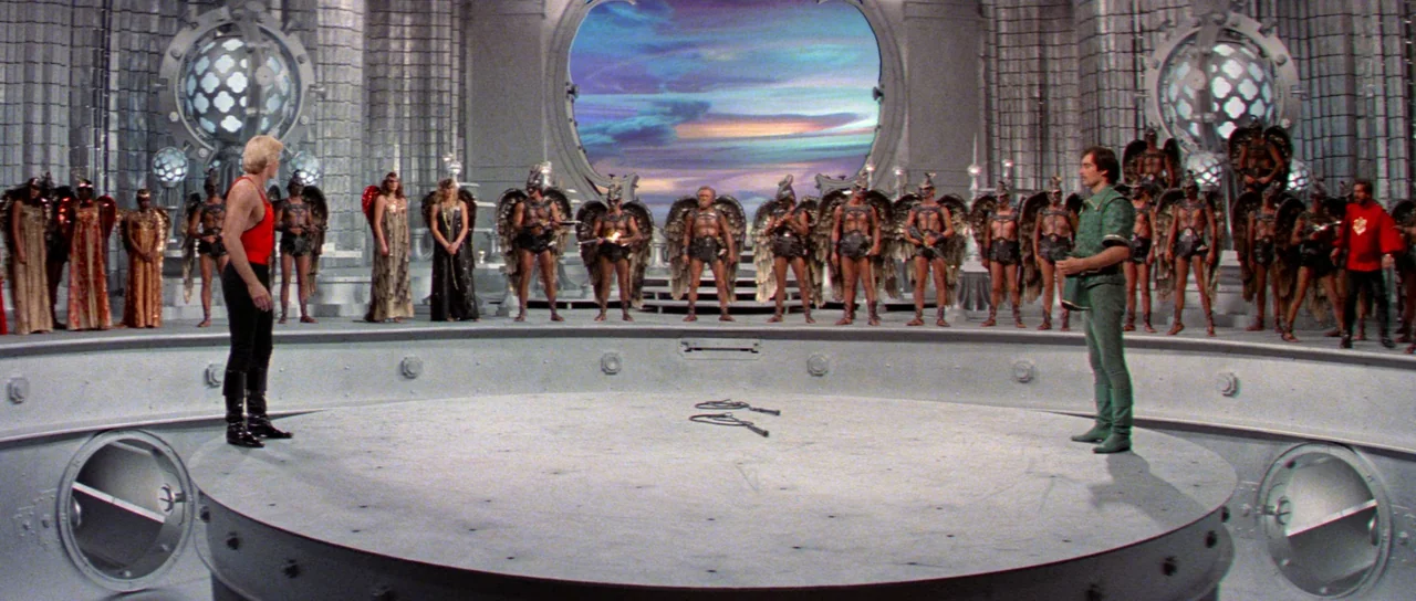Flash Gordon (1980) movie still: wide — A massive, circular metallic arena features decorative sphere structures and a rear circular window. Two men…