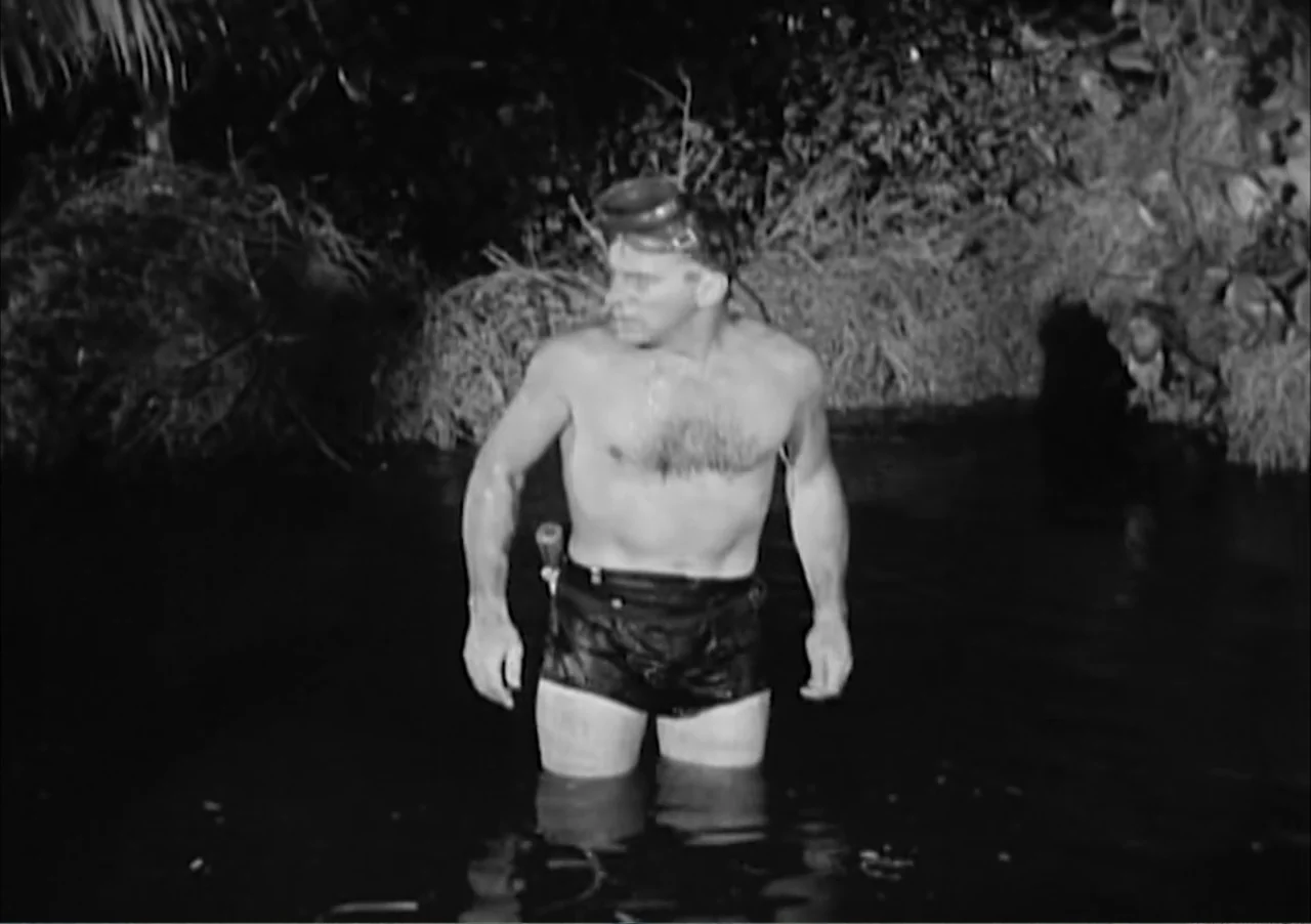 Creature from the Black Lagoon (1954) movie still: medium — A dark river bank at night is surrounded by dense, overgrown vegetation. A shirtless man…