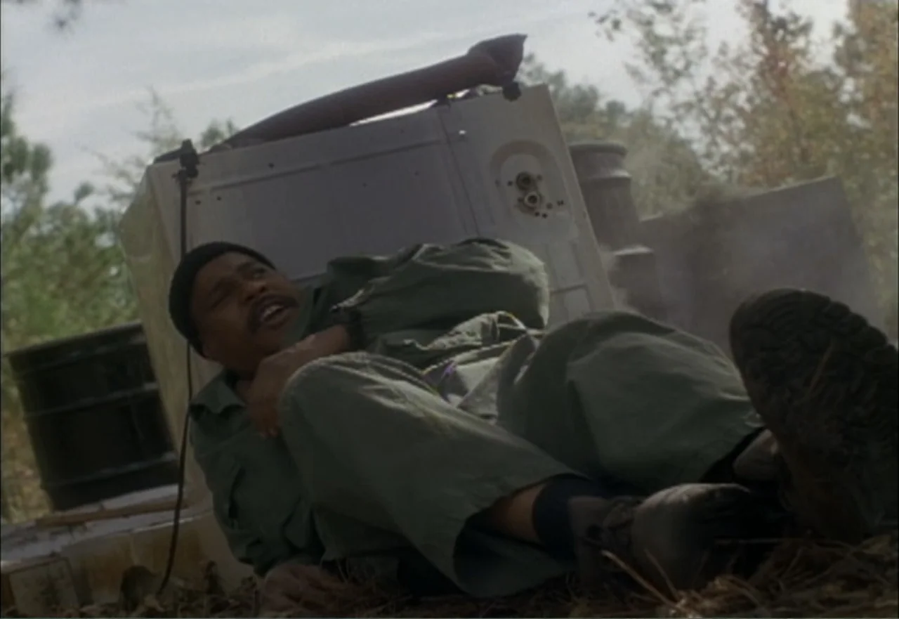 Ambushed (1998) movie still: medium — An outdoor junkyard area with scattered debris and a large discarded white appliance. A man lies on the ground…