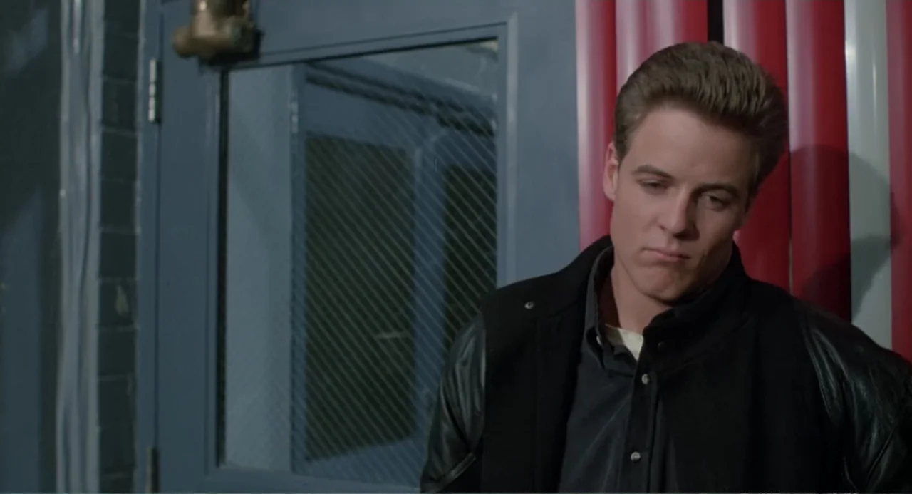 Dangerously Close (1986) movie still: medium — A doorway interior features a dark blue metal door with a glass pane. A young man with dark hair…