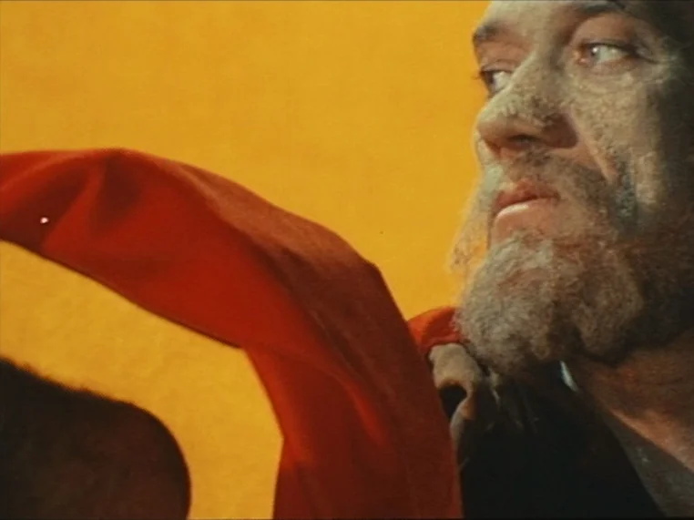 Salome (1972) movie still: closeup — A man stands in front of a flat, solid yellow background. He looks toward the left while a large red cloth…