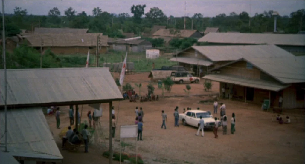 The Killing Fields (1984) movie still: establishing — A rural camp composed of simple wooden structures with corrugated metal roofs sits on dirt.…