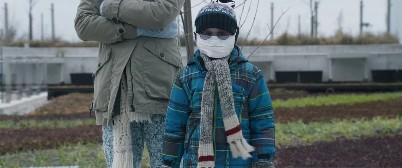 Room (2015) movie still: medium — A rooftop planting area with rows of small crops under an overcast sky. A child in a plaid jacket and face mask…