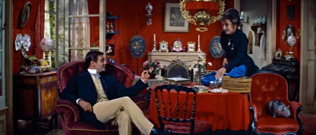 Gigi (1958) movie still: wide — A lavish Victorian parlor features deep red walls, ornate furniture, and numerous decorative ornaments. A man seated…