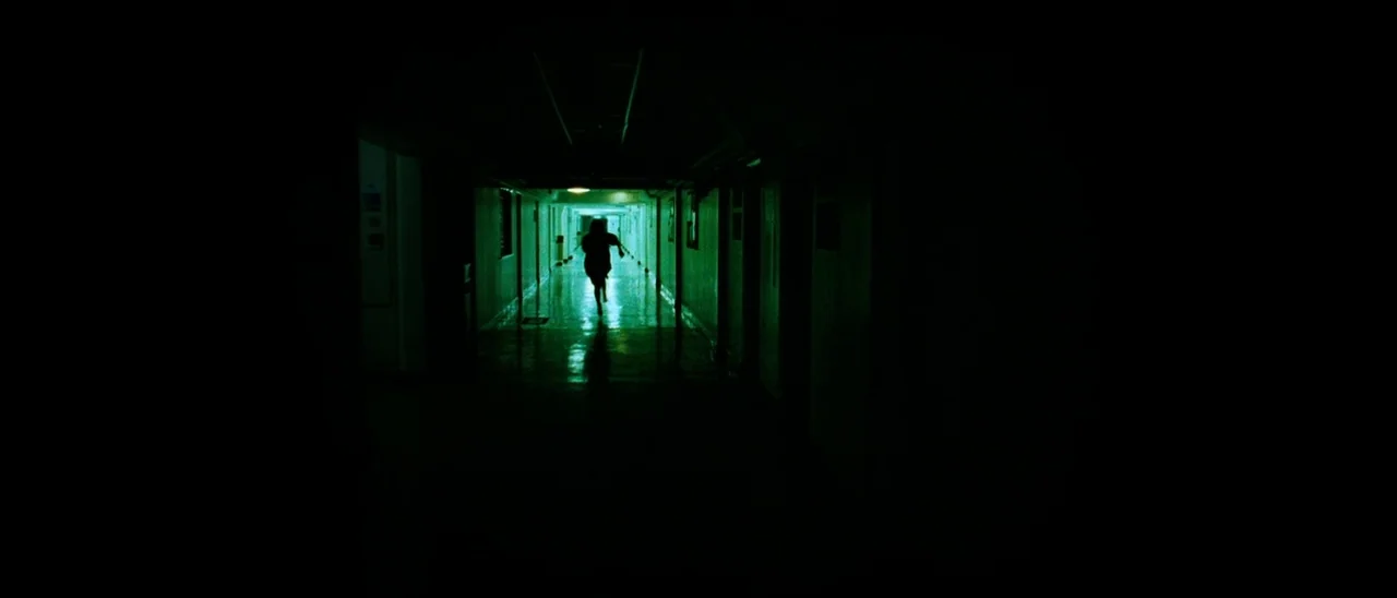 The Descent (2005) movie still: wide — A long, dimly lit institutional hallway features reflective flooring and deep surrounding darkness. A…