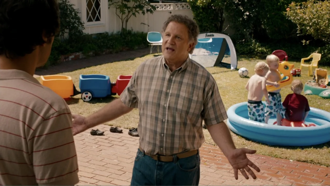This Is 40 (2012) movie still: medium — A residential backyard features a brick patio area and a grass lawn with toys. A middle-aged man stands on…