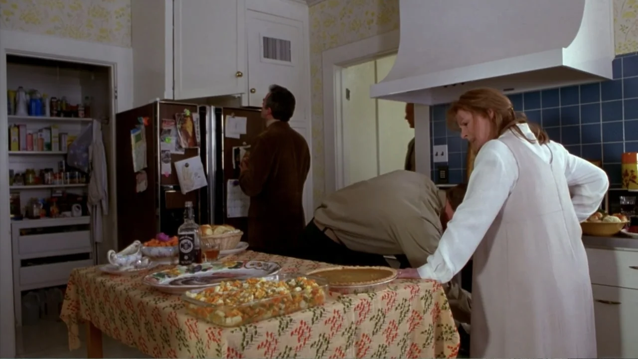 Music from Another Room (1998) movie still: medium — A suburban home interior features a dining table covered in food and floral wallpaper. A woman…