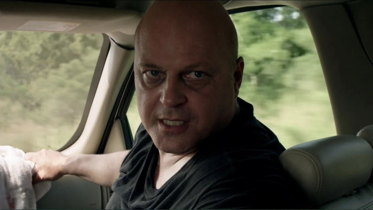 Parker (2013) movie still: medium — The interior of a moving car shows a man in the passenger seat. The man stares directly at the viewer as the…