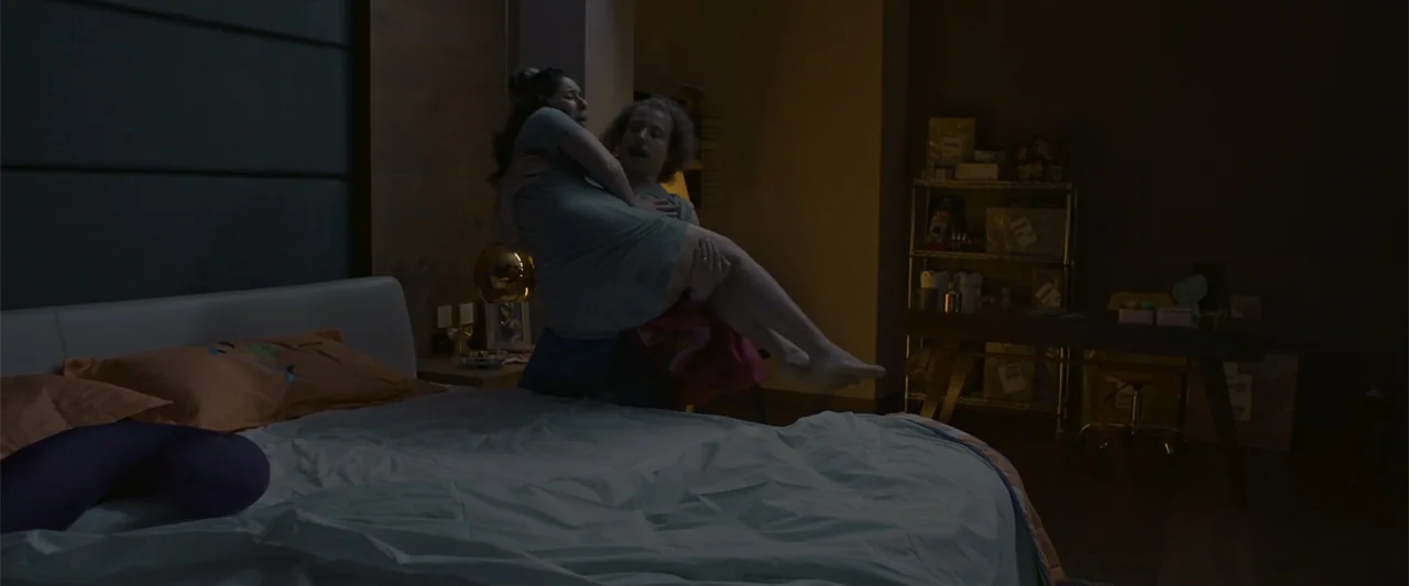 Mirreyes contra Godínez 2: El retiro (2022) movie still: medium — A darkened room features a bed in the foreground and a desk in the back. A man…