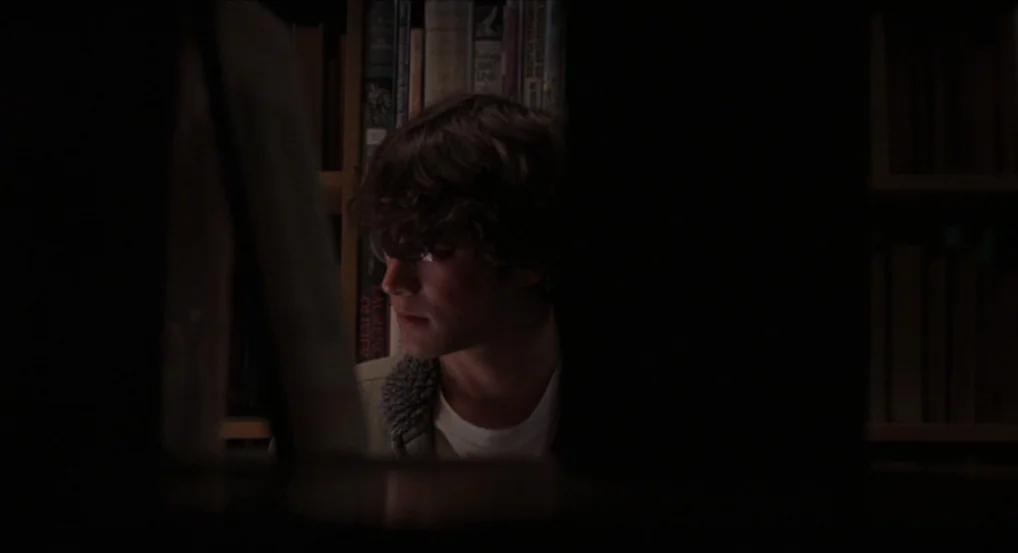 Brick (2005) movie still: closeup — A dimly lit home library features shelves filled with books in the background. A young man sits in the dark…