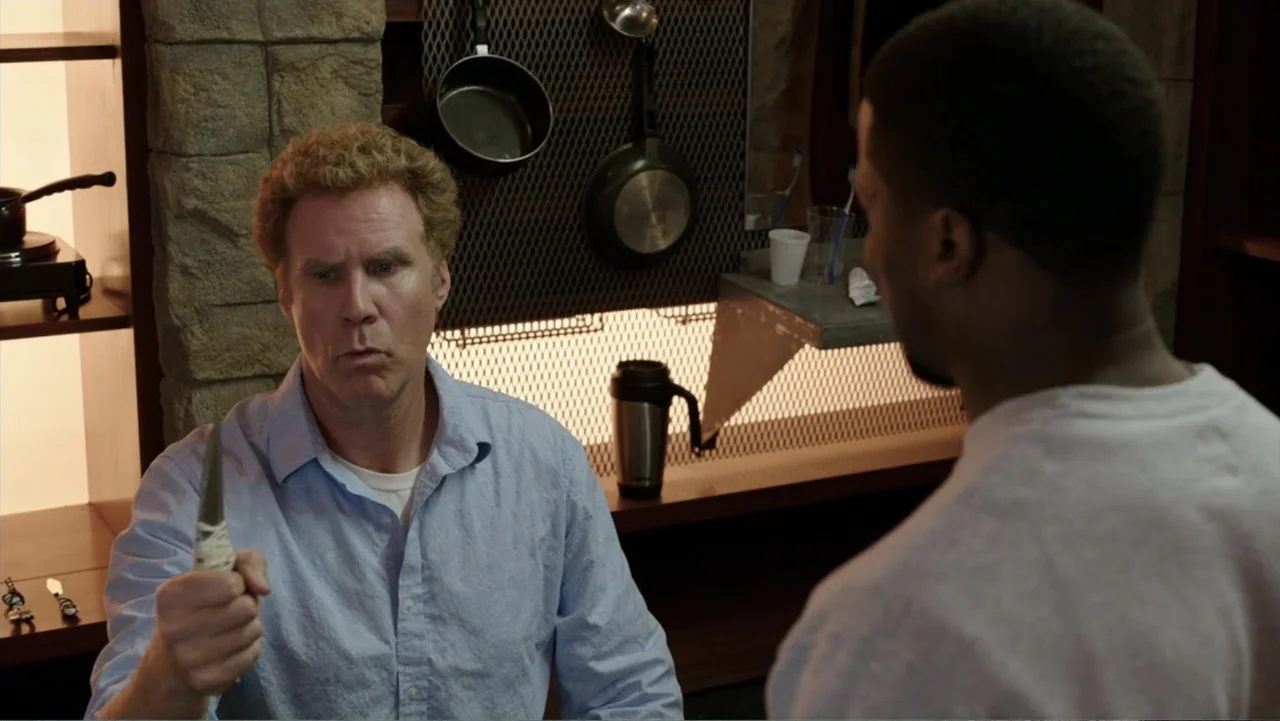 Get Hard (2015) movie still: medium — A spartan room features stone walls, hanging cookware, and a desk with personal items. A man in a blue shirt…