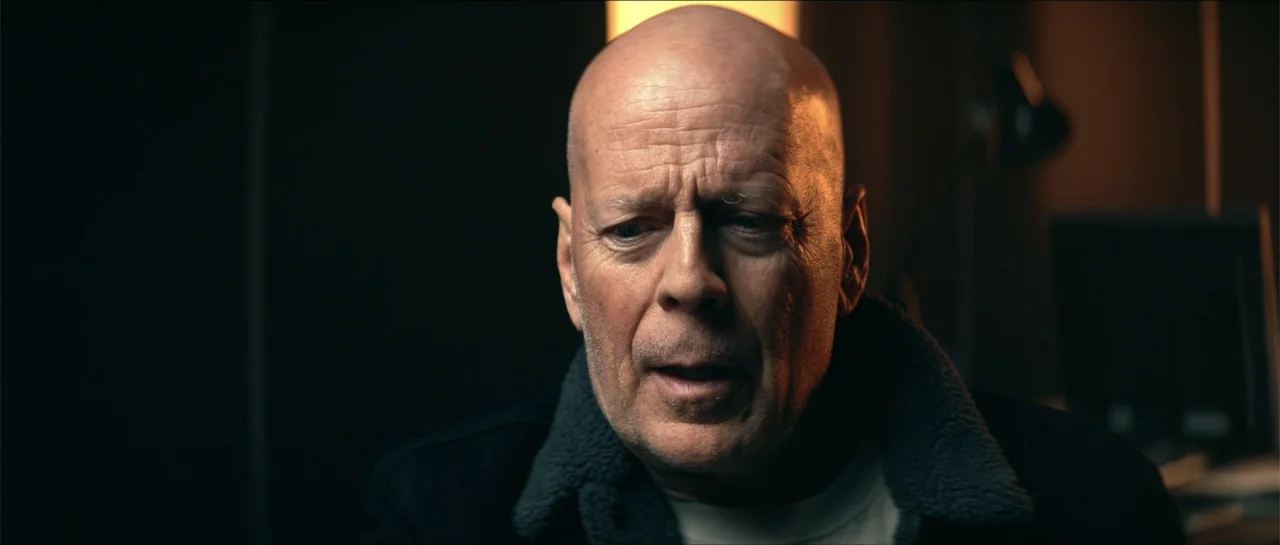 Cosmic Sin (2021) movie still: closeup — A dimly lit indoor space with shadows obscuring the surroundings. A bald older man in a jacket looks…