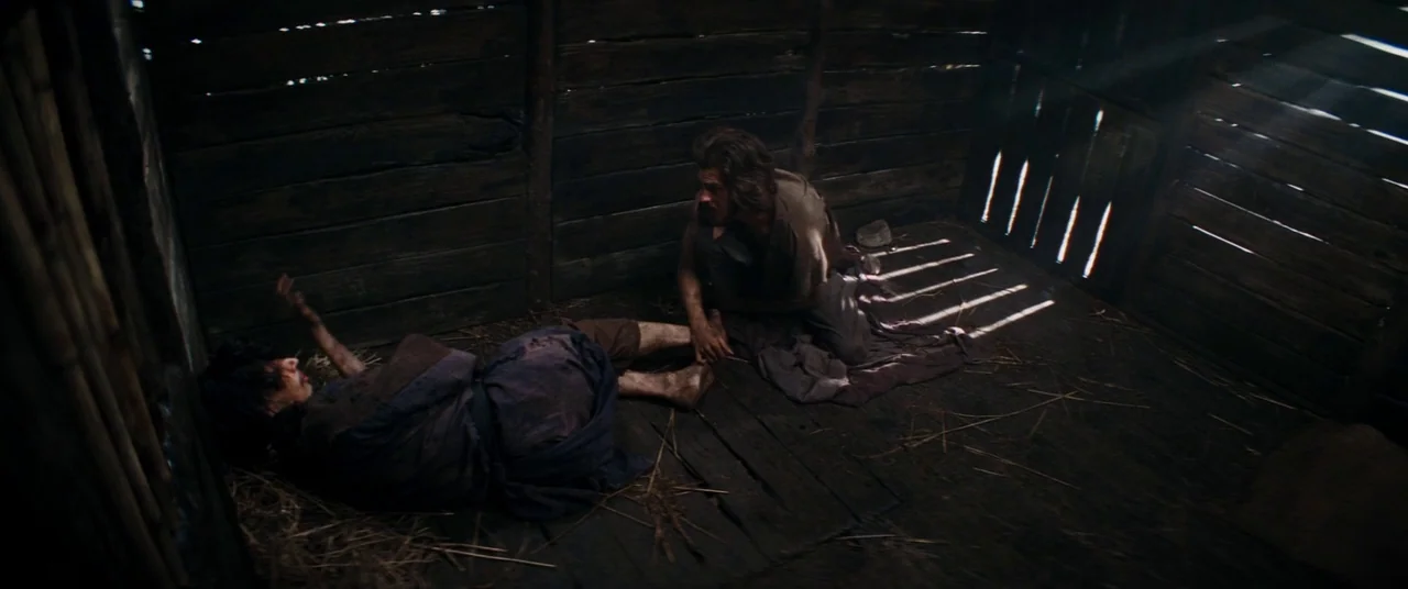 Silence (2016) movie still: wide — A dimly lit cabin features dark wooden plank walls and a straw-strewn floor. One man sits on the floor watching…
