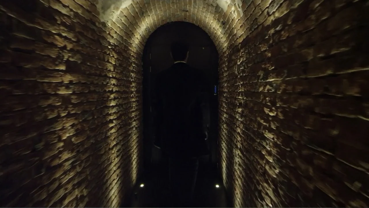 Entre las sombras (2025) movie still: medium — A narrow arched hallway made of dark red bricks with dim recessed floor lighting. A man in a suit…