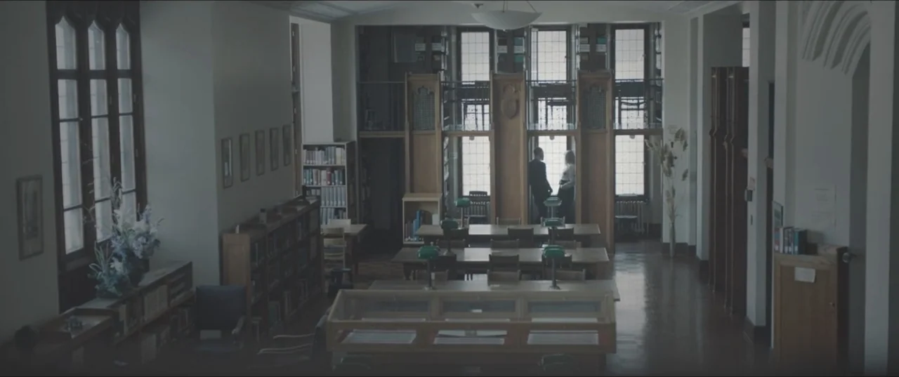 Zoe (2018) movie still: wide — A spacious library contains rows of wooden desks and tall, shelving-lined windows. A man and a woman stand in the…