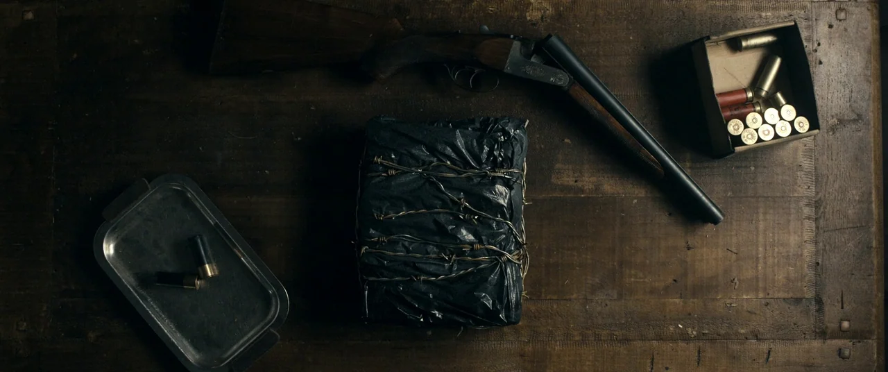 Evil Dead (2013) movie still: wide — A dark wooden tabletop displays a collection of weapons and mysterious wrapped items. A black package wrapped…