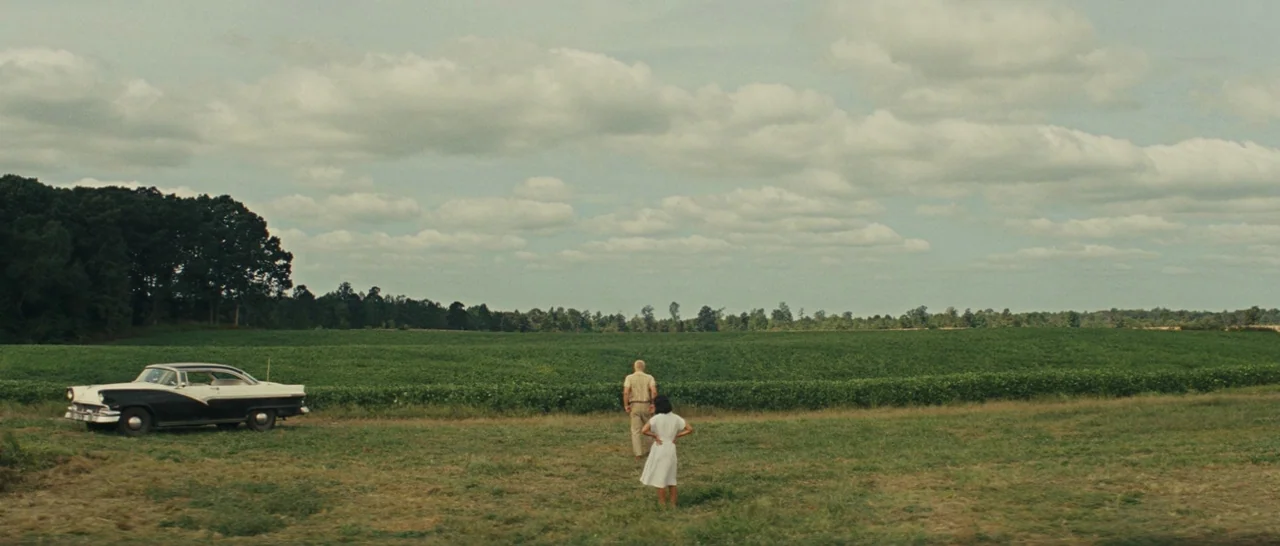 Loving (2016) movie still: wide — An open agricultural field stretches toward a treeline under a large cloudy sky. A man and a woman stand near a…