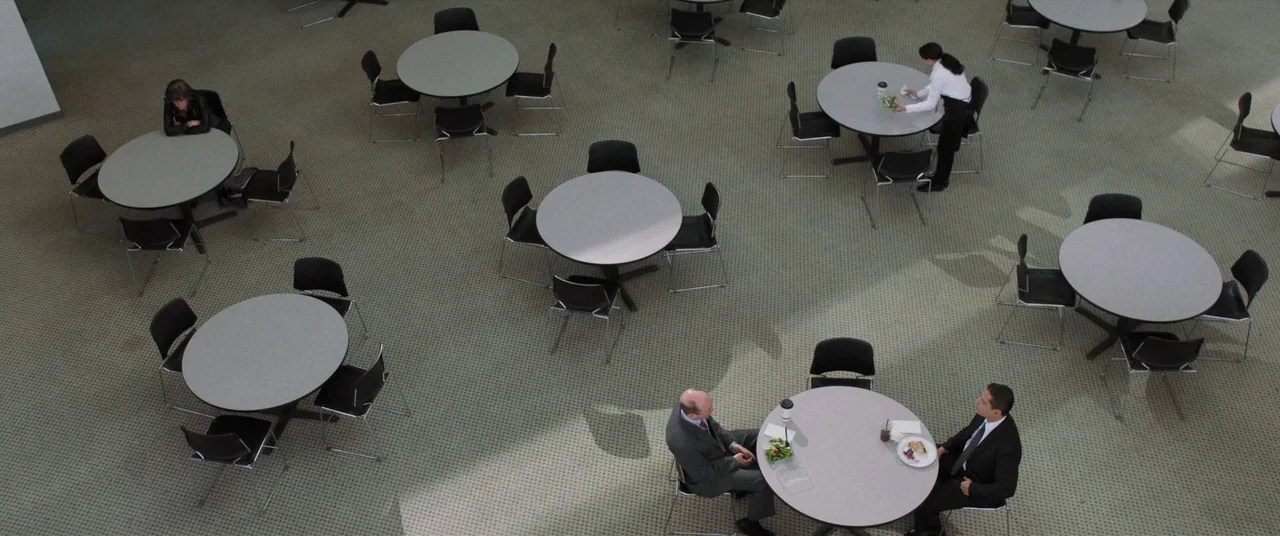 Destroyer (2018) movie still: wide — A spacious cafeteria features light grey circular tables arranged across a patterned carpet floor. Two men in…