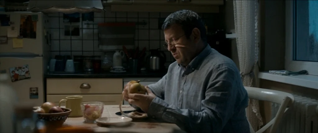 Graduation (2016) movie still: medium — A dimly lit kitchen features tiled walls, a refrigerator, and a wooden table. A man in a blue shirt sits at…
