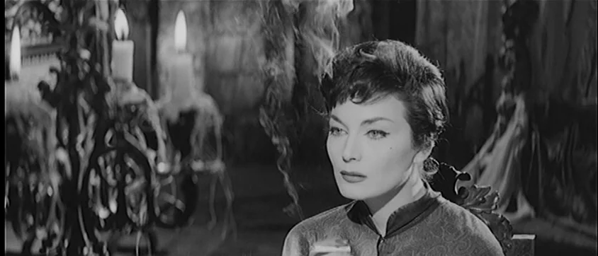 I Vampiri (1957) movie still: closeup — An interior stone room features a candelabra with flickering flames in the foreground. A woman with dark…