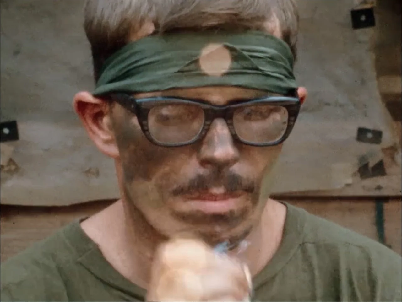 One to One: John & Yoko (2025) movie still: closeup — A man in a military-style shirt and headband stands outdoors in a camp area. The man looks…