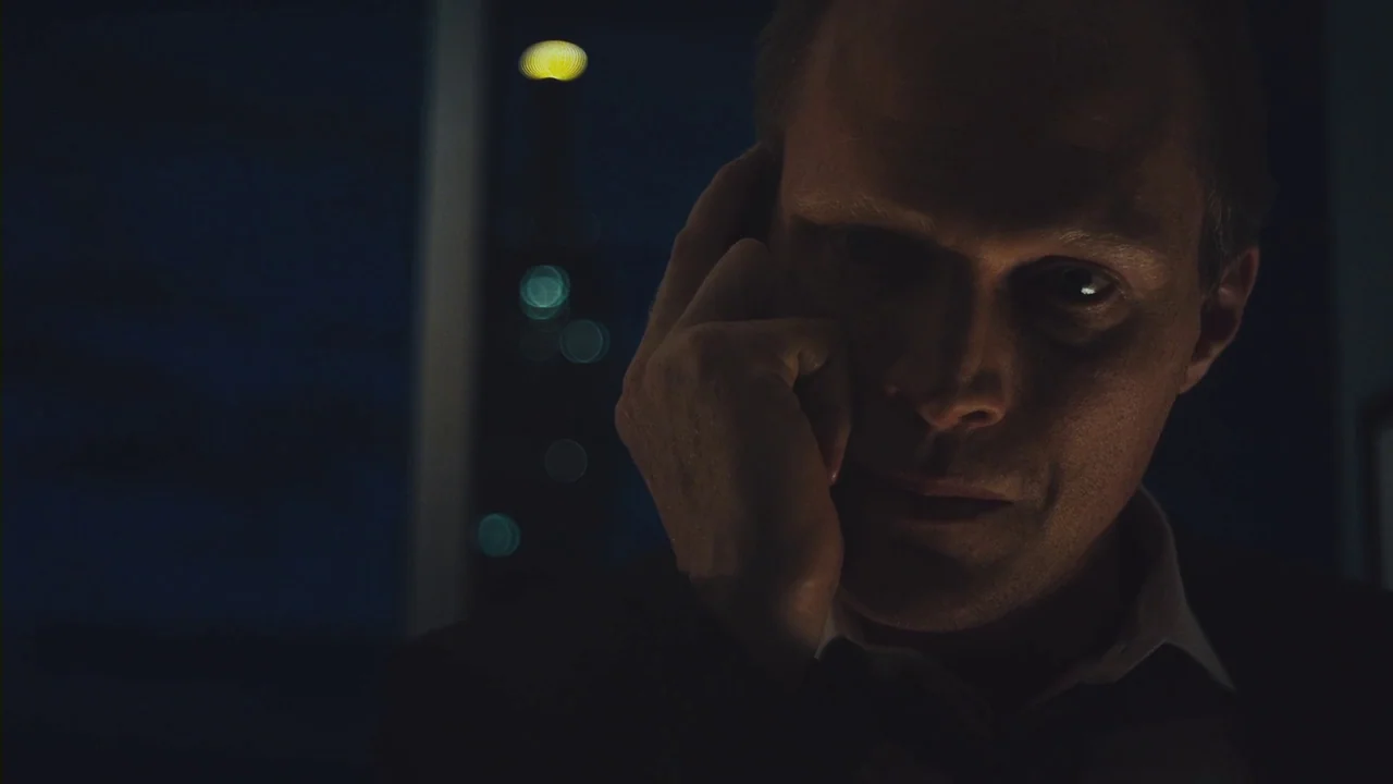 Margin Call (2011) movie still: closeup — A dimly lit high-rise office at night with city lights blurred in the background. A man sits in darkness…