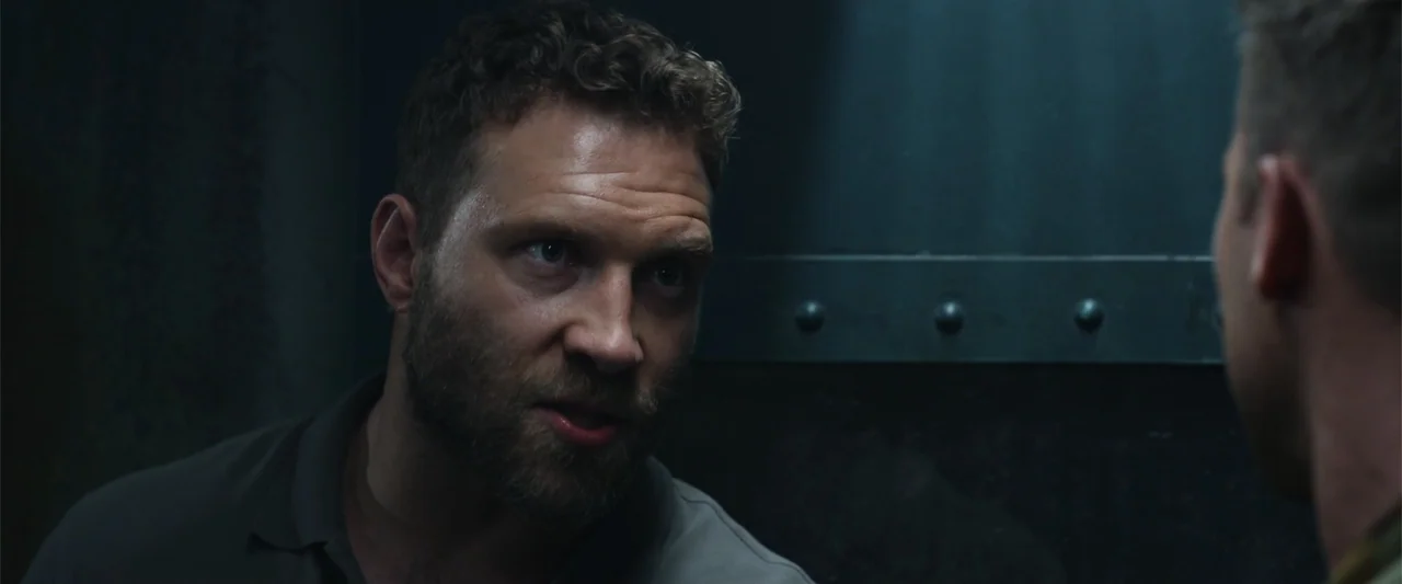 Black Site (2022) movie still: closeup — A dimly lit interior room features a dark industrial metal wall with rivets. A man in a grey polo shirt…