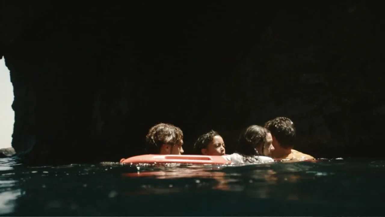 Jurassic World Rebirth (2025) movie still: medium — Four people swim together in the dark water within a large limestone sea cave. They tread water…