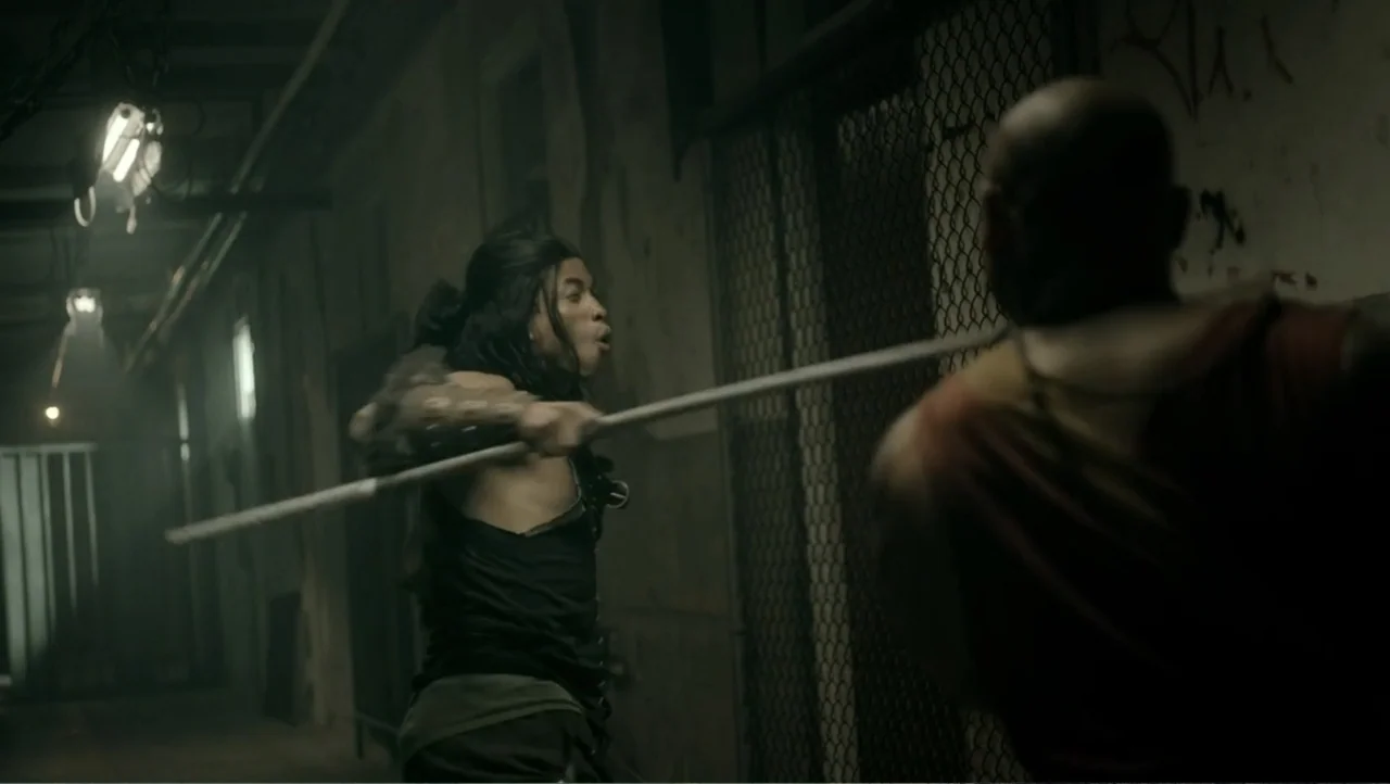 Shadow Master (2022) movie still: medium — A dimly lit industrial corridor with chain-link fencing and concrete walls. A man holds a long staff out…